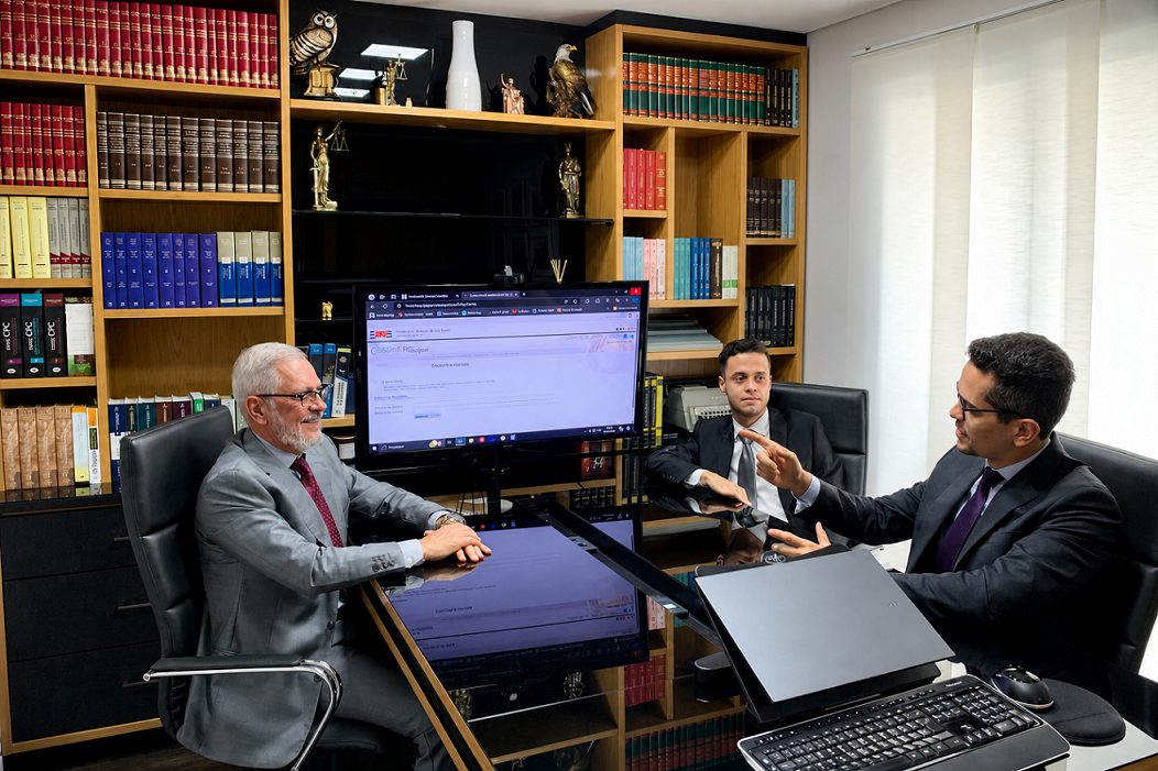 Os advogados Sérgio Lucio Ruffo, Danilo Roberto Fernandes e Lucas Leal Leite, da esquerda para a direita, conversando em torno da mesa de reunião do escritório, diante de uma estante de livros jurídicos