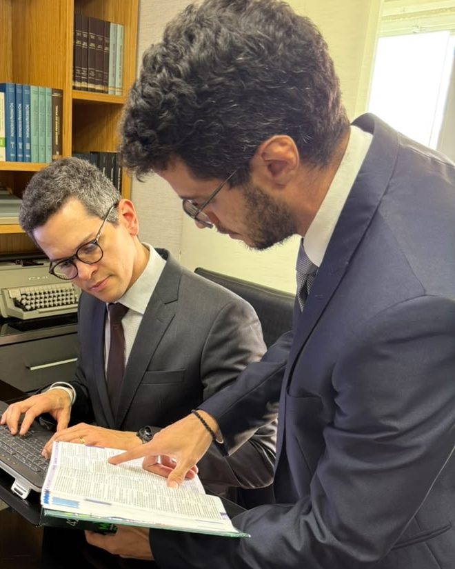 Os advogados Lucas Leal Leite, sentado, e Danilo Roberto Fernandes, em pé, consultam juntos um código de leis, diante da estante do escritório
