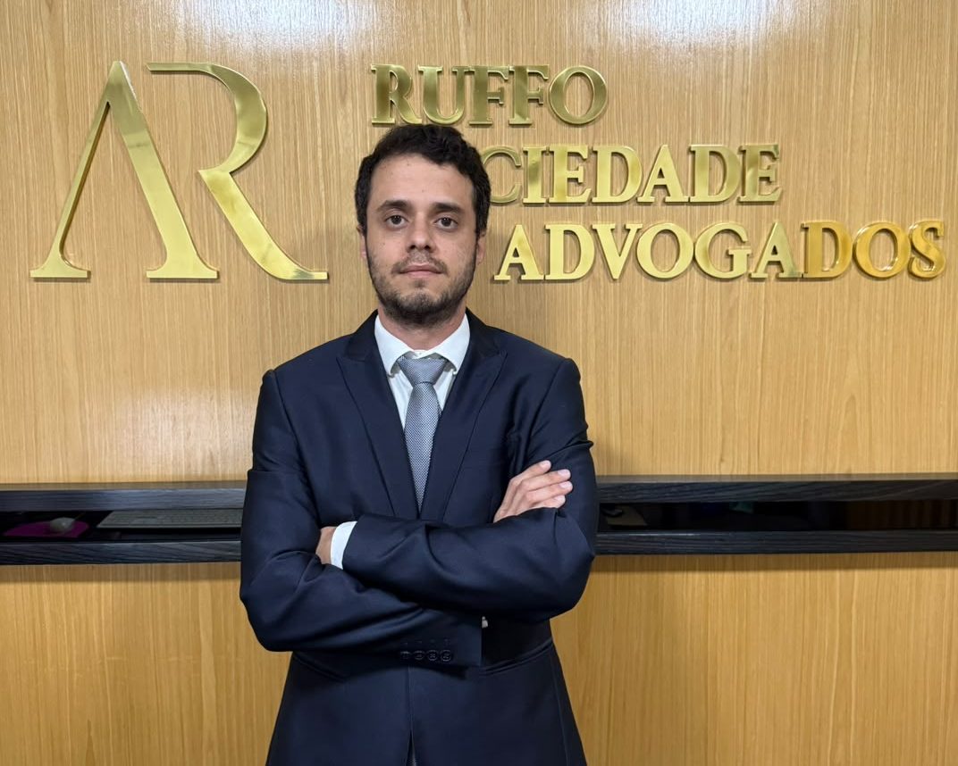 Danilo Roberto Fernandes, de terno e braços cruzados, diante do letreiro dourado com o nome da Ruffo Sociedade de Advogados, na recepção do escritório
